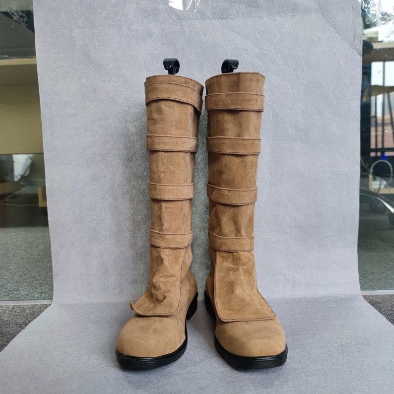 K&ouml;nnte beinhalten: Ein Paar braune Wildleder-Stiefel, die bis zum Knie reichen, mit mehreren Riemen und Schnallen. Die Stiefel haben eine spitze Zehenpartie und einen niedrigen Absatz.