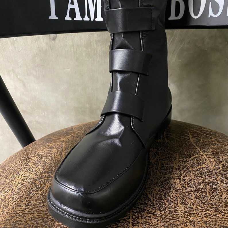 May include: Black leather boots with a buckle strap and a pointed toe. The boots are sitting on a brown textured surface.