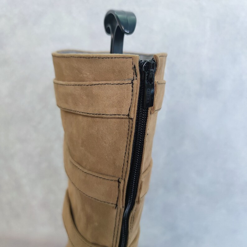 K&ouml;nnte beinhalten: Beigefarbene, kniehohe Stiefel aus Wildleder mit einem schwarzen Rei&szlig;verschluss auf der R&uuml;ckseite. Die Stiefel haben mehrere Riemen und Schnallen.