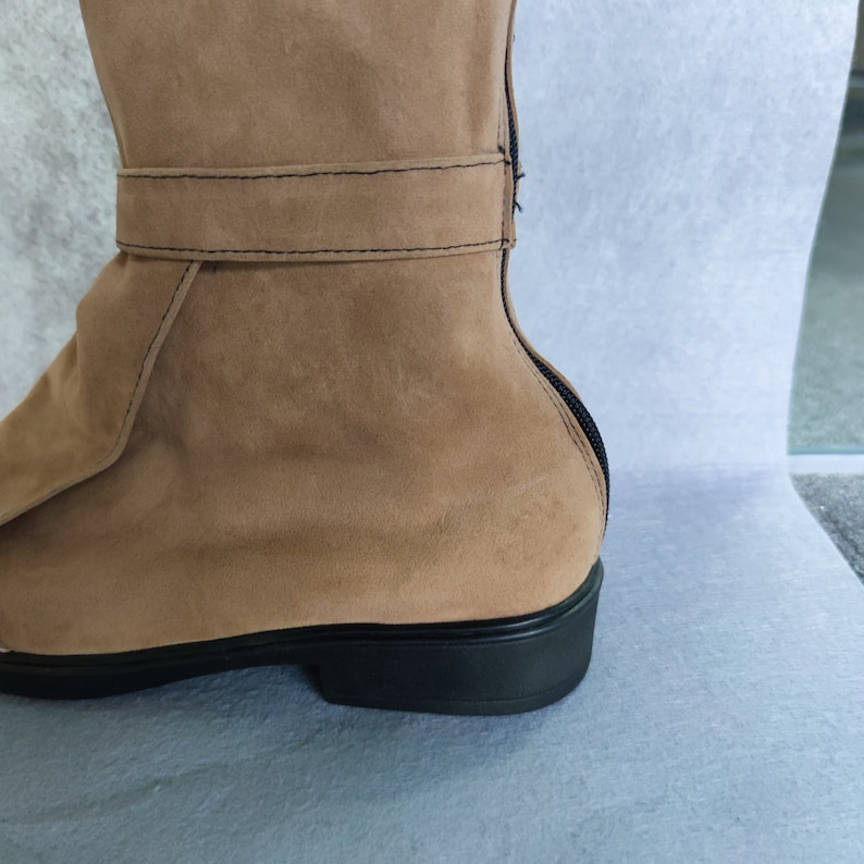 K&ouml;nnte beinhalten: Ein brauner Wildlederstiefel mit schwarzem Rei&szlig;verschluss und schwarzer Sohle. Der Stiefel hat einen umgeschlagenen Schaft und eine Schnalle.