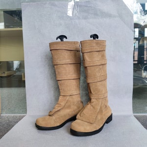 K&ouml;nnte beinhalten: Ein Paar braune Wildlederstiefel, die bis zum Knie reichen, mit mehreren Riemen und Schnallen. Die Stiefel haben eine spitze Zehenpartie und einen niedrigen Absatz.