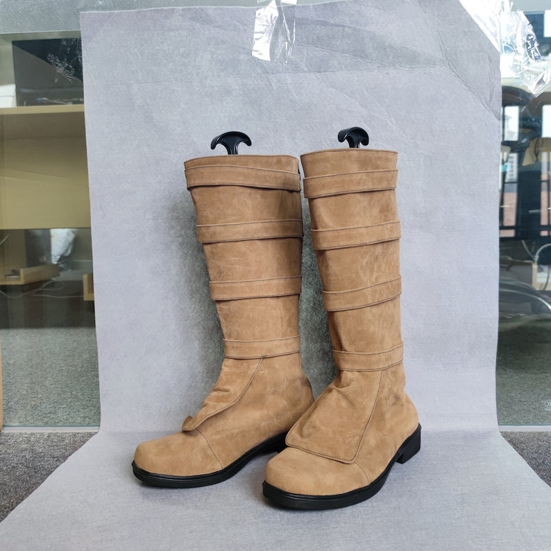 K&ouml;nnte beinhalten: Ein Paar braune Wildlederstiefel, die bis zum Knie reichen, mit mehreren Riemen und Schnallen. Die Stiefel haben eine spitze Zehenpartie und einen niedrigen Absatz.