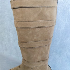 K&ouml;nnte beinhalten: Ein Paar braune, kniehohe Stiefel aus Wildleder mit mehreren Riemen und N&auml;hten.