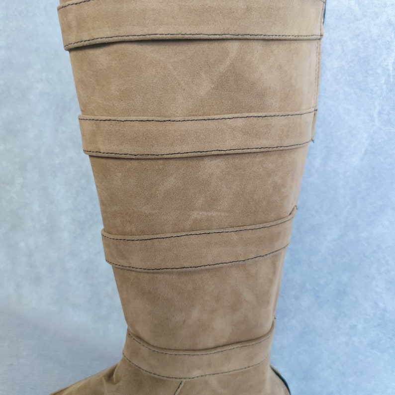 K&ouml;nnte beinhalten: Ein Paar braune, kniehohe Stiefel aus Wildleder mit mehreren Riemen und N&auml;hten.