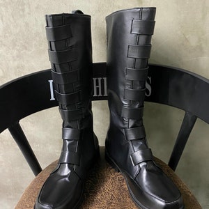 Puede incluir: Un par de botas negras de cuero de caña alta con un detalle de hebilla en la parte posterior del eje. Las botas tienen una punta puntiaguda y un tacón bajo.