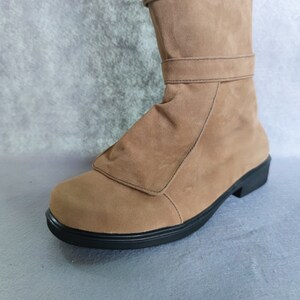 K&ouml;nnte beinhalten: Ein Paar braune Wildlederstiefel mit schwarzer Sohle. Die Stiefel haben ein Umlege-Design am oberen Rand und einen einzigen Riemen &uuml;ber dem Fu&szlig;r&uuml;cken.