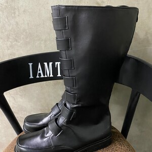 May include: Black leather boots with a buckle strap design. The boots have a tall shaft and a rounded toe.