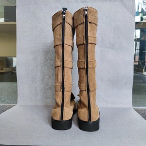 K&ouml;nnte beinhalten: Ein Paar braune, kniehohe Stiefel aus Wildleder mit mehreren Schnallen und einem Rei&szlig;verschluss auf der R&uuml;ckseite.