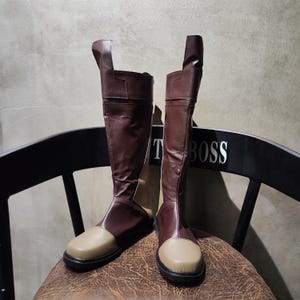 Puede incluir: Un par de botas altas de cuero marrón con punteras de color beige. Las botas están sobre una silla de madera con el texto "T BOSS" en el respaldo.