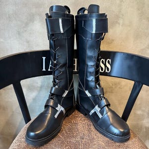 May include: Black knee-high boots with silver detailing. The boots have a lace-up front, adjustable straps, and a flat sole. The boots are shown on a chair, with the letters "IA" and "SS" visible on the backrest.