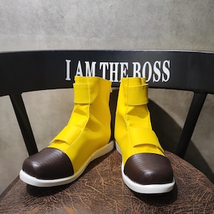 Puede incluir: Un par de botines amarillos con punteras de cuero marrón y suelas blancas. Las botas tienen una correa en la parte superior del pie y están sentadas en una silla de madera con las palabras "I AM THE BOSS" impresas en el respaldo.