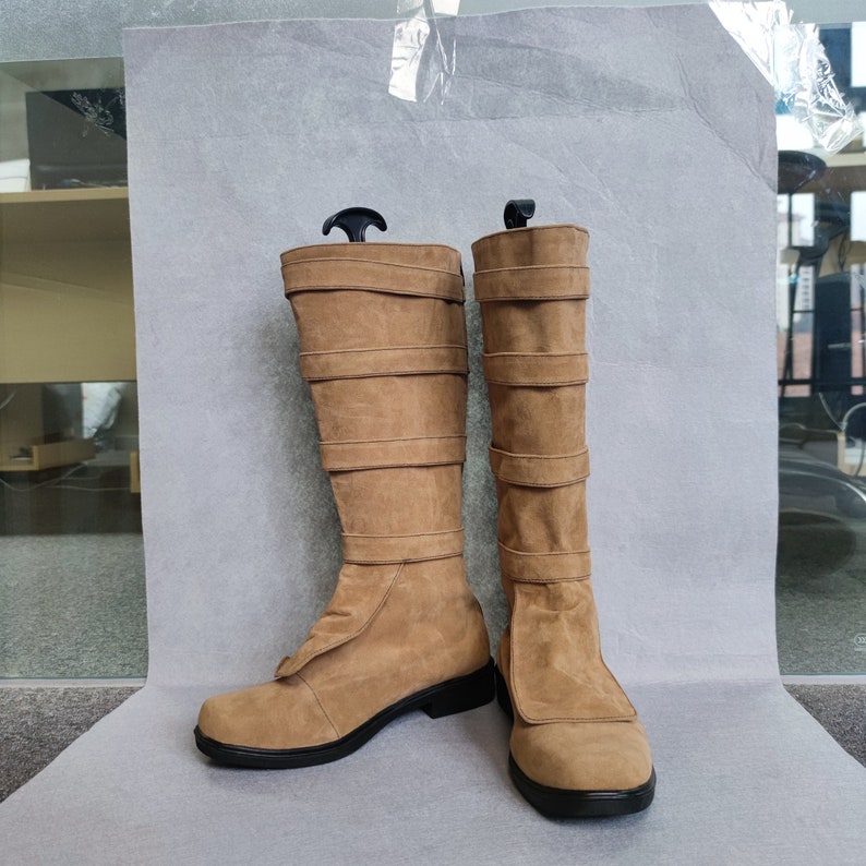 K&ouml;nnte beinhalten: Ein Paar braune, kniehohe Stiefel aus Wildleder mit schwarzen Schnallen und einer schwarzen Sohle.