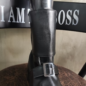 Op de afbeelding: Zwarte leren enkellaarsjes met een gespsluiting en een omslag aan de bovenkant. De laarsjes hebben een bord met de tekst "I AM THE BOSS" op de achtergrond.