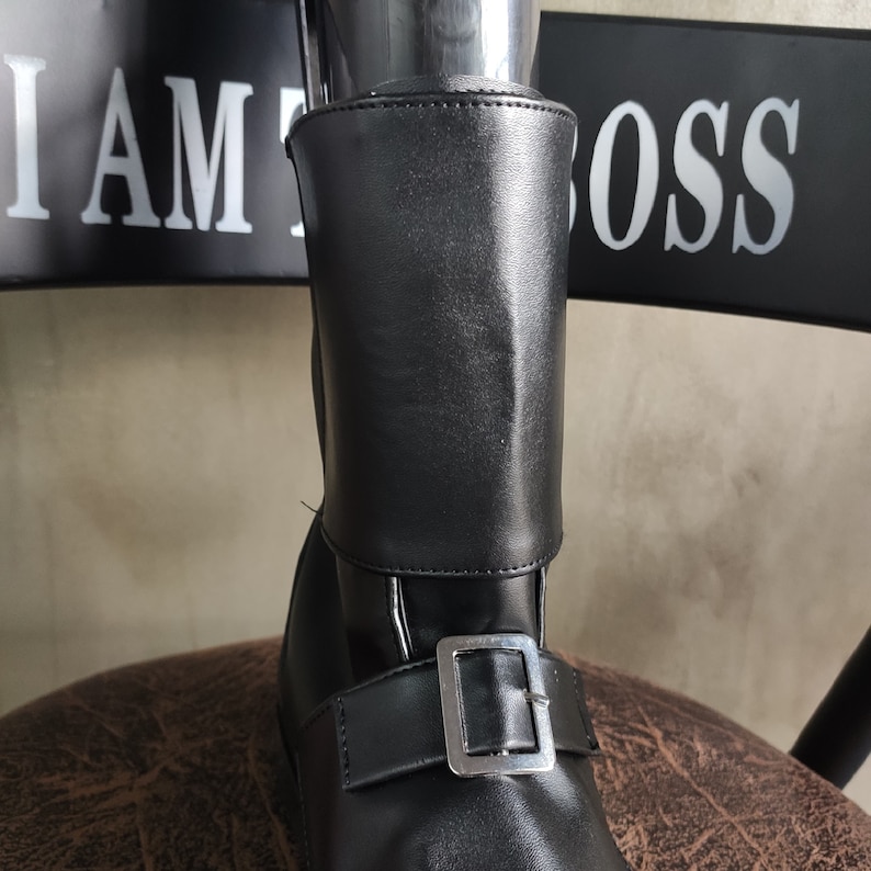 Op de afbeelding: Zwarte leren enkellaarsjes met een gespsluiting en een omslag aan de bovenkant. De laarsjes hebben een bord met de tekst "I AM THE BOSS" op de achtergrond.