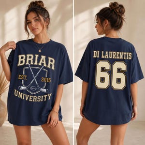 Op de afbeelding: Marineblauw T-shirt met de tekst "BRIAR UNIVERSITY" en een afbeelding van gekruiste hockeysticks op de voorkant. De achterkant toont "DI LAURENTIS" en het nummer "66" in crème. Het shirt heeft een casual, oversized pasvorm.