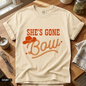 Puede incluir: Camiseta de color marfil con el texto "SHE'S GONE BOW" en naranja. El diseño incluye un sombrero de vaquero y letras estilo cuerda. La camiseta está hecha de un material suave y tiene mangas enrolladas.