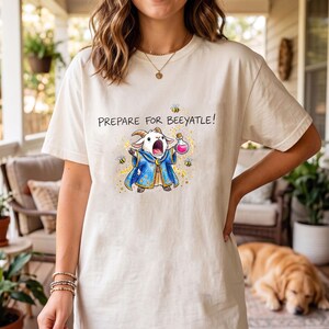 Può includere: T-shirt color crema con un cartone animato di una capra vestita da mago, che tiene in mano una boccetta di pozione. Il testo "PREPARE FOR BEYATLE!" è sopra. La capra è circondata da api e scintille. Un capo d'abbigliamento casual.