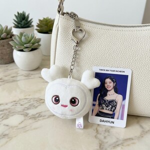 May include: A white plush keychain in the shape of a cartoon character with heart-shaped ears and pink accents, attached to a white purse. A photo card with the text "TWICE 6TH TOUR INCHEON" and "DAHYUN" is also visible.