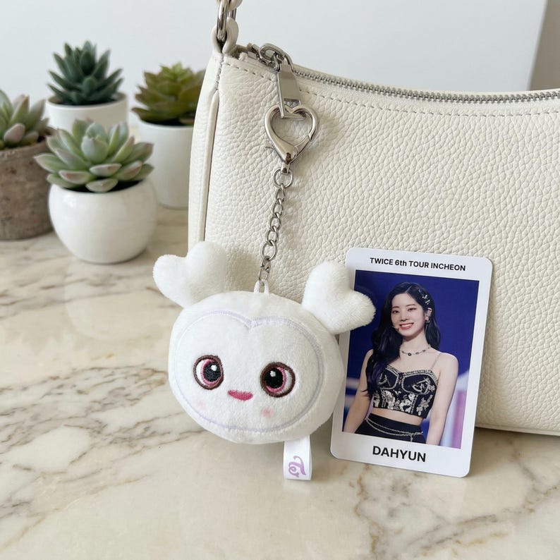 May include: A white plush keychain in the shape of a cartoon character with heart-shaped ears and pink accents, attached to a white purse. A photo card with the text "TWICE 6TH TOUR INCHEON" and "DAHYUN" is also visible.