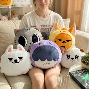 Puede incluir: Una colección de cojines de felpa con diseños de personajes de animales y dibujos animados. Los cojines incluyen un gato gris, un conejito blanco, un pollito amarillo, un personaje morado y un oso blanco. Los cojines son suaves y achuchables.