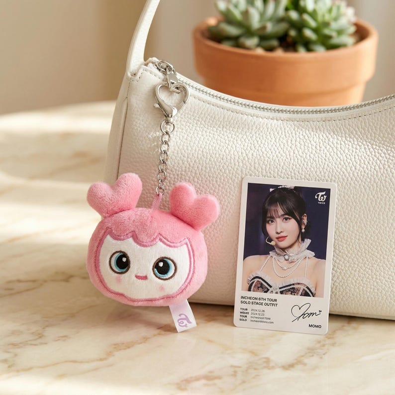 May include: A pink plush keychain shaped like a cartoon character with heart-shaped ears, attached to a white purse. A photo card featuring a woman in a stage outfit is next to the keychain. The keychain has a tag with a logo.