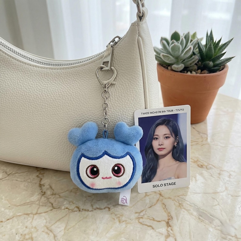 May include: A light blue plush keychain in the shape of a cartoon character with heart-shaped ears, attached to a silver chain. A photo card with the text "TWICE INCHEON 6TH TOUR - TZUYU" is next to the keychain. A white purse is in the background.