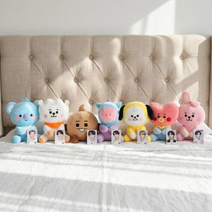 May include: A collection of seven plush character dolls in various colors, including blue, white, brown, purple, yellow, and pink. Each doll has a unique design and is accompanied by a small card. The dolls are arranged on a white bed.