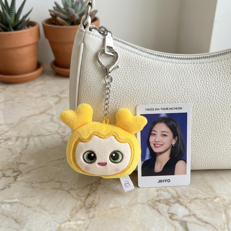 May include: A yellow plush keychain in the shape of a cartoon character with heart-shaped ears, attached to a silver chain. A photo card with the text "TWICE 6th TOUR INCHEON" and the name "JIHYO" is next to the keychain. The keychain is hanging from a white purse.
