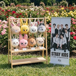 Może przedstawiać: Drewniany stojak wystawowy prezentuje pluszowe plecaki w kształcie zwierząt w różnych kolorach, w tym białym, różowym i żółtym. W pobliżu znajduje się baner z grupą osób i napisem "STRAY KIDS". Tło wypełniają kolorowe kwiaty.