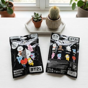 May include: Two black BT21 Backpack Buddies blind bags are displayed on a white surface. The bags feature colorful cartoon characters and the text "BT21." The bags are sealed and ready to collect. A small succulent, cactus, and other plants are in the background.