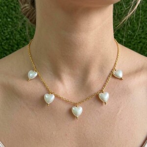May include: A gold chain necklace with six white heart-shaped pendants. Each heart has a small gold bead. The necklace is worn around the neck.