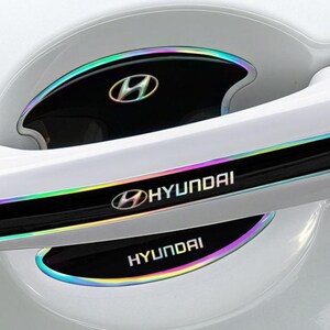 May include: Car door handle trim in white, with iridescent detailing. The trim displays the Hyundai logo and name in white. The design incorporates a black base with a rainbow-effect border, enhancing the vehicle's exterior appearance.