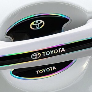 May include: Car door handle accents with the Toyota logo and text. The black accents have a rainbow iridescent trim. The Toyota logo is in silver. The car door is white.
