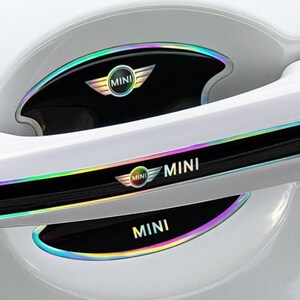 May include: Car door handle accents with a black base and iridescent trim. The accents feature the MINI logo and text in a silver color. The car door is white.
