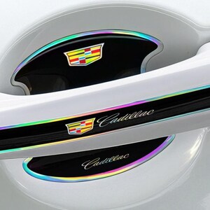May include: White car door handle accents with iridescent trim. Features the Cadillac logo and script in silver. The accents are black with a colorful, rainbow-like border, enhancing the car's exterior.