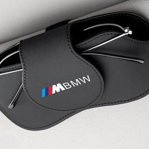 Puede incluir: Estuche de gafas de cuero negro con el logotipo de BMW en blanco, azul y rojo. El estuche está abierto, revelando un par de gafas con montura plateada. El estuche está diseñado para guardar y proteger las gafas.