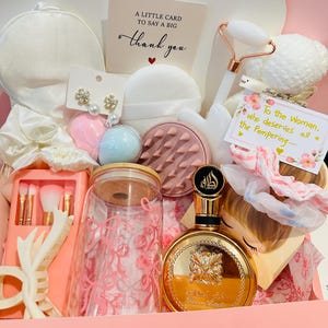 Op de afbeelding: Een roze geschenkdoos gevuld met schoonheids- en zelfzorgartikelen. Inclusief een gouden parfumfles, een gezichtsroller, make-up borstels en een kaart met "thank you". De doos is versierd met roze en witte accenten.