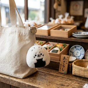 Puede incluir: Un llavero de peluche blanco de onigiri (bola de arroz) con una cara linda y detalles de algas negras, colgando de un bolso de mano de lona. El fondo muestra una estanter&iacute;a de madera con comida y vajilla japonesa.