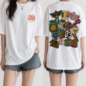 May include: White t-shirt with a graphic design on the front and back. The front features the text "Noah Kahan" in red and the back has a collage of colorful patches with text and illustrations, including "American Cars" and "23".
