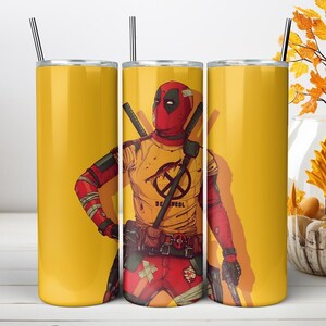 May include: A cylindrical stainless steel tumbler with a yellow background featuring a graphic illustration of the character Deadpool wearing a red mask and a yellow t-shirt with a black logo. The tumbler includes a metal straw and a lid.