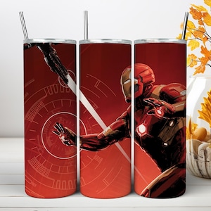 May include: A cylindrical stainless steel tumbler featuring a red and gold Iron Man character graphic with a glowing chest arc reactor and palm repulsor. The design includes a circular technical HUD pattern and a thin metal straw at the top.