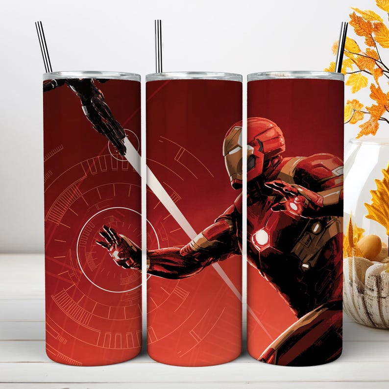 May include: A cylindrical stainless steel tumbler featuring a red and gold Iron Man character graphic with a glowing chest arc reactor and palm repulsor. The design includes a circular technical HUD pattern and a thin metal straw at the top.