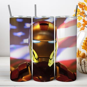May include: A cylindrical metal tumbler featuring a graphic of a red and gold armored helmet with glowing blue eyes. The background displays a blurred American flag pattern. The tumbler includes a thin metal straw inserted into the top lid.