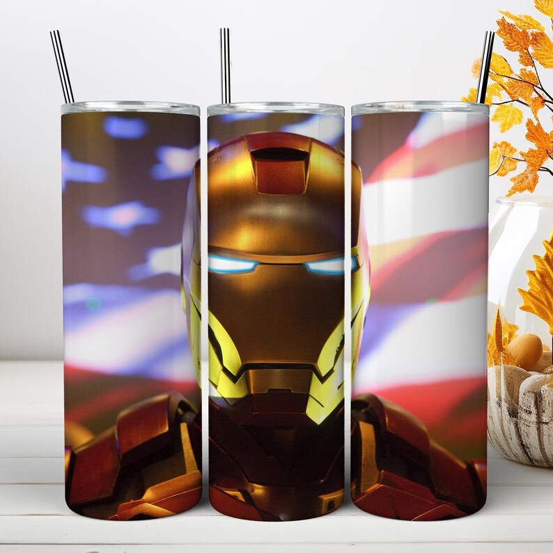 May include: A cylindrical metal tumbler featuring a graphic of a red and gold armored helmet with glowing blue eyes. The background displays a blurred American flag pattern. The tumbler includes a thin metal straw inserted into the top lid.