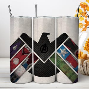 May include: Three cylindrical tumblers with metal straws, featuring a white background and a large black eagle silhouette. The design incorporates geometric panels with red, blue, green, and purple colors, including a central Avengers logo.