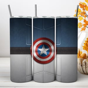 May include: A cylindrical stainless steel tumbler featuring a graphic design inspired by a superhero shield. The design includes a central red, white, and blue star emblem, a dark blue upper section, a grey lower section, and a black horizontal band.