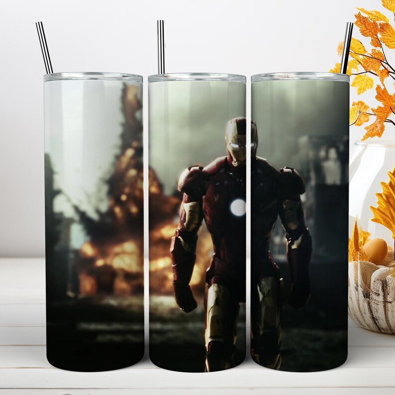 May include: A cylindrical stainless steel tumbler featuring a graphic print of a red and gold armored superhero standing in front of a fiery explosion. The tumbler includes a clear lid and a thin metal straw, showcasing a detailed action scene.