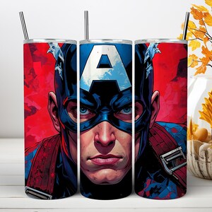 May include: A cylindrical tumbler featuring a graphic illustration of Captain America with a prominent blue mask, a white letter A on the forehead, and a red background. The tumbler includes a thin metal straw extending from the top opening.