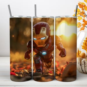 May include: A cylindrical metal tumbler featuring a graphic of a red and gold armored character in a crouched position. The background displays a blurred autumn forest scene with golden sunlight filtering through the trees. A thin metal straw is included.