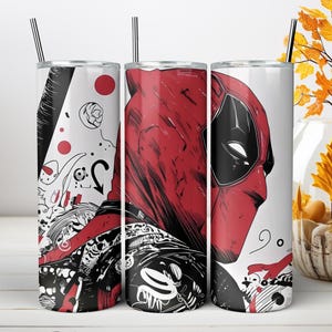 May include: A cylindrical stainless steel tumbler featuring a graphic illustration of a red and black masked character. The design includes abstract black and white line art, red circles, and a metal straw inserted into the top lid of the container.